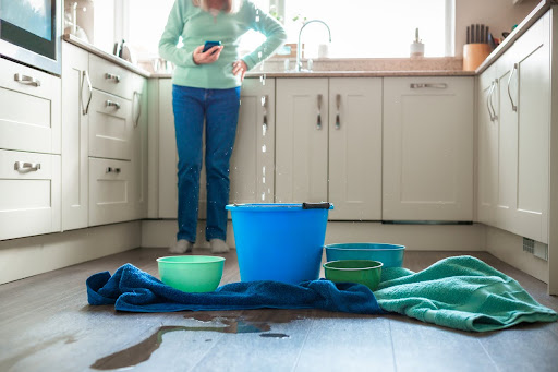 Worried woman with buckets and towels under a leaking kitchen ceiling, calling for help.